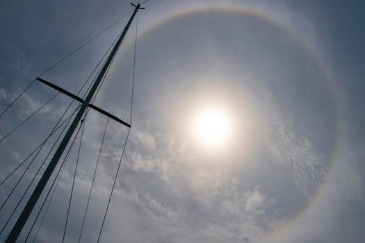 A perfect circle of rainbow-coloured light framed the sun in an atmospheric phenomenon, with the silhouette of the mast of yacht visible with sheets. Photo location: Cres Island, Croatia