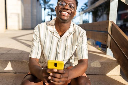 Happy black man sitting on stairs, relaxing outdoors using mobile phone, laughing. Lifestyle concept.