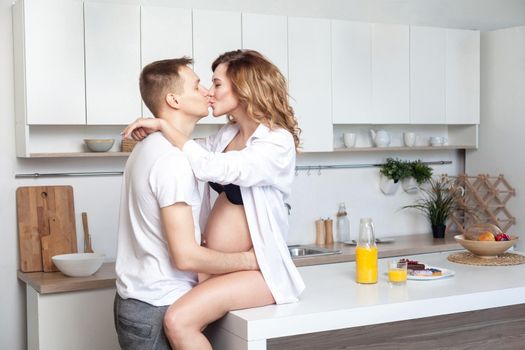 Lovestory. Young married couple embraces siting on table in kitchen. Husband hugs his pregnant wife, putting his hands on her big belly, parents enjoy each other and kissing with closed eyes.