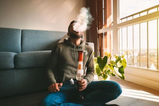 Man smoking bong and exhaling the smoke at home. Man smoking pot, medical marijuana or cannabis from a bong or water pipe. Cannabis and weed legalisation concept. High quality photo