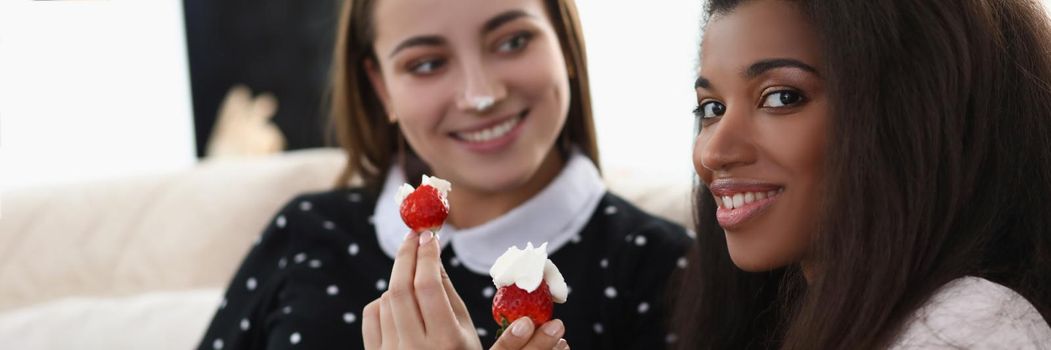 Portrait of smiling girls eating tasty fresh strawberry covered with whipped cream. Romantic date at home, best friends chilling. Lgbt, bff, lunch concept
