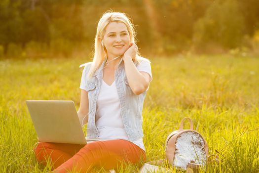 Young woman with laptop sits on the grass in the park on a sunny day. Laughing blonde in a white tank top and shorts. Online training, remote work and communication in social networks.