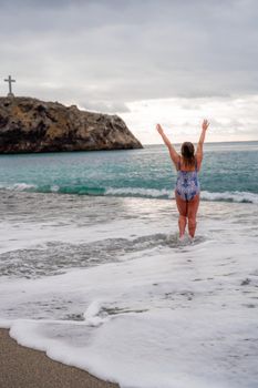 A plump woman in a bathing suit enters the water during the surf. Alone on the beach, Gray sky in the clouds, swimming in winter