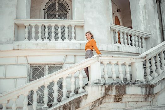 A middle-aged woman looks like a good blonde with curly beautiful hair and makeup on the background of the building. She is wearing a yellow sweater