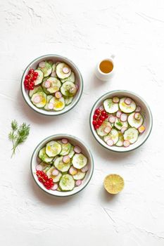 radish and cucumber salad served in three round bowls decorated with red currants, white concrete background, top view