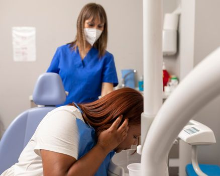 A black woman patient is spitting out after rinsing her mouth while waiting for the dentist to start the treatment at the dental clinic