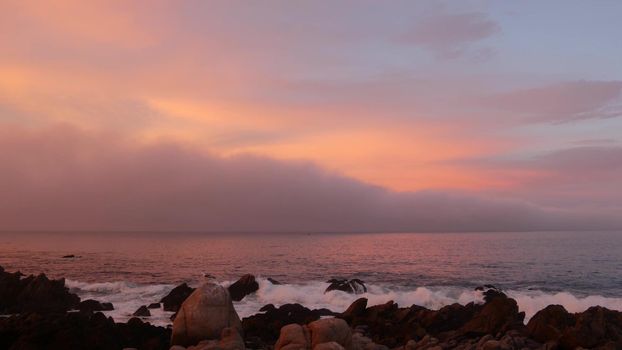 Rocky craggy ocean beach, calm sea waves, Monterey bay, 17-mile drive seascape, California coast, USA. Beachfront waterfront Pacific Grove, waterside promenade. Pink purple pastel dramatic sunset sky.