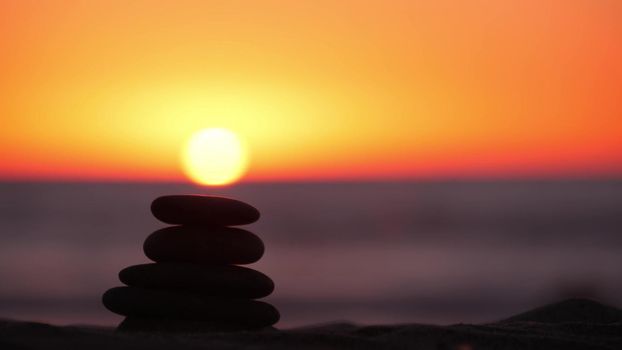 Stack of pebble stones, sandy ocean beach, sunset sky. Rock balancing in sun light, sea water waves. Stones staking in pyramid pile. Zen meditation and harmony in balance. Calm tranquil atmosphere.