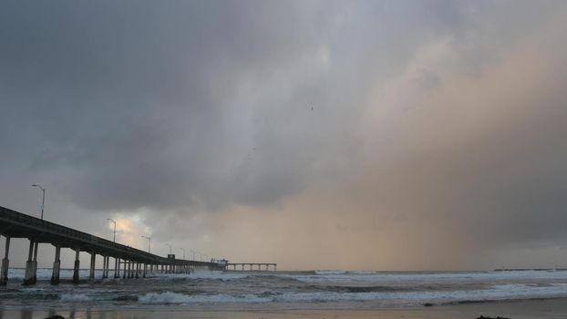 Ocean Beach pier in rainy weather, sea waves in foggy air, California coast, USA. Beachfront boardwalk on piles in water, San Diego. Misty cloudy dramatic shore under pouring downpour. Railfall drops.