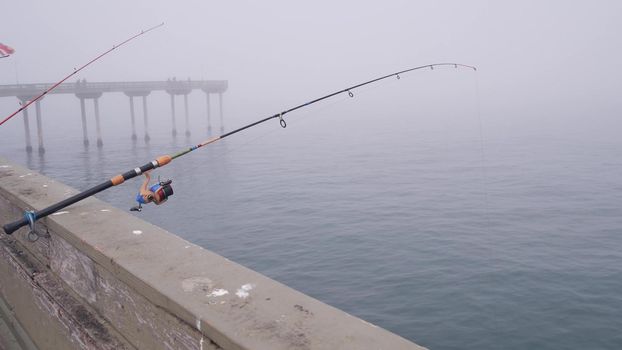 Wooden Ocean Beach pier in fog, misty California coast, USA. Foggy moody cloudy weather on San Diego shore. Calm tranquil silent atmosphere. Waterfront boardwalk. Fishing or recreational angling rod.