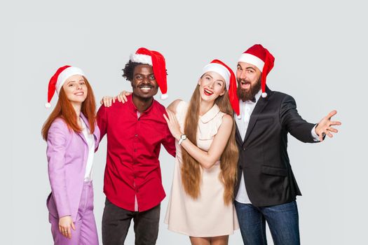 Group of beautiful happy handsome friends in red cap standing and celebrating new year, looking at camera with toothy smile. indoor studio shot, isolated on gray background.