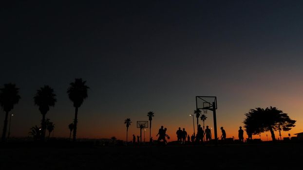 Silhouettes of players on basketball court outdoor, people playing basket ball game, sunset ocean beach, California coast, USA. Black hoop, net and backboard on streetball sport field. Mission beach.