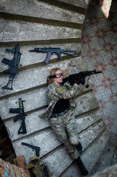 Caucasian woman in military uniform lies on the stairs of an abandoned building and holds a machine gun. View from above