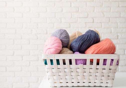 pastel colored yarn wool in a white basket on white brick wall background, front view. Hobbies and leisure. Interior details