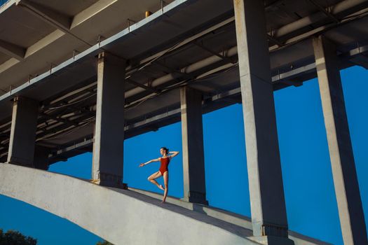 Beautiful young strong healthy woman ballerina gymnast in red leotard posing as model on bridge girder and blue sky and looking into the distance.