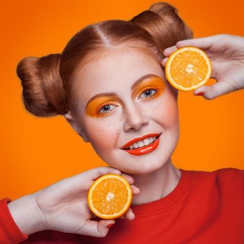 Young beautiful funny fashion model with orange slice on orange background. with orange makeup and hairstyle and freckles. studio shot, looking at camera with smile.