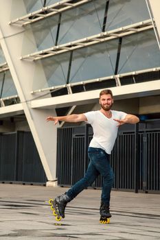 Profesional Bearded man on rollerblades standing in building background. Young fit man with white t-shirts and jeans on roller skates riding outdoors after rain.