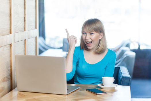 Have idea! Portrait of creative positive attractive young girl freelancer in blue t-shirt are sitting in cafe and working on laptop with toothy smile and showing finger up, looking at camera. indoor