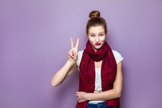 Portrait of beautiful young happy smiling woman, showing two fingers or victory gesture, dressed white t-shirt and red scarf on purple background.
