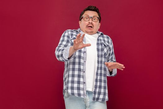 Portrait of shocked handsome middle aged business man in casual checkered shirt and eyeglasses standing and looking at camera with scared eyes. indoor studio shot, isolated on dark red background.