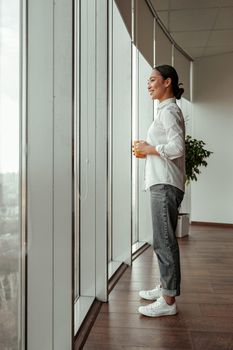 Smiling asian female worker drinking coffee standing near windows at office. Business concept