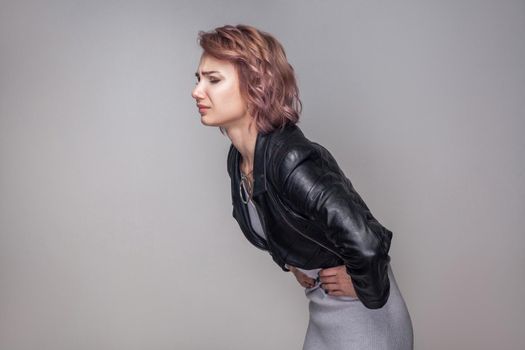 Stomach pain. Profile side view portrait of woman with short hairstyle in casual style black leather jacket standing and holding her sick belly. indoor studio shot, isolated on grey background.