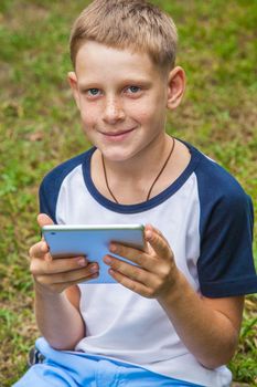 Cute young caucasian kid with freckles on his face in blue shorts and white and blue T-shirt and tablet and working in park and smiling. top view, looking at camera.