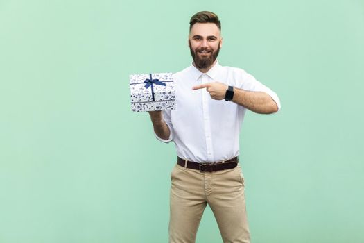 Young adult businessman pointing finger on gift box and looking at camera and toothy smile. On light green background. Studio shot
