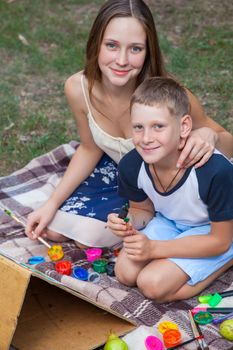 Older sister trying to teach her brother to paint and posing in the park on summer time, looking at camera and smiling. top view.