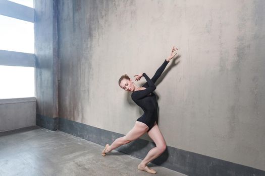 Ballerina movement in classic ballet posing near window. Studio shot, gray wall
