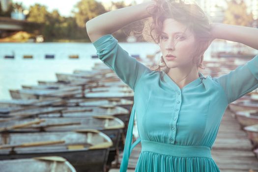 Beautiful young fashion model in green turquois dress and fashion makeup and hairstyle is posing on pier, posing and looking at camera. close up. sunset effect on camera. with passion.