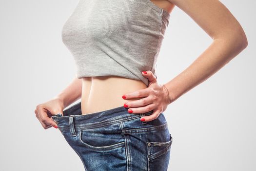 Closeup of slim waist of young woman standing in big jeans and gray top showing successful weight loss, indoor studio shot, isolated on light gray background, diet concept.