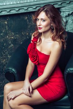 Beautiful young fashion model in red dress and fashion makeup and hairstyle posing sitting in the armchair near the artificial fireplace and looking at camera. Studio shot.