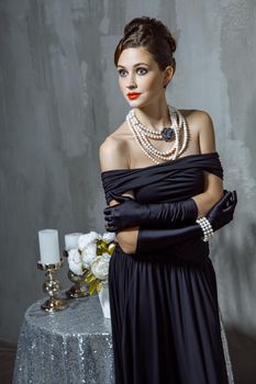 Beautiful young fashion caucasian model in black long dress and fashion makeup and beautiful hairdo posing sitting on the chair and looking at camera. Studio shot.