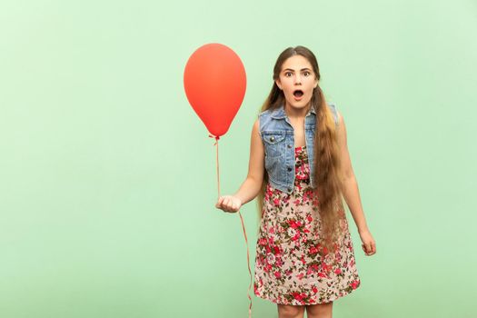 Unbelievable! The beautiful blonde teenager is sho?ked, looking at camera, with red ballon on a light green background. Isolated studio shot