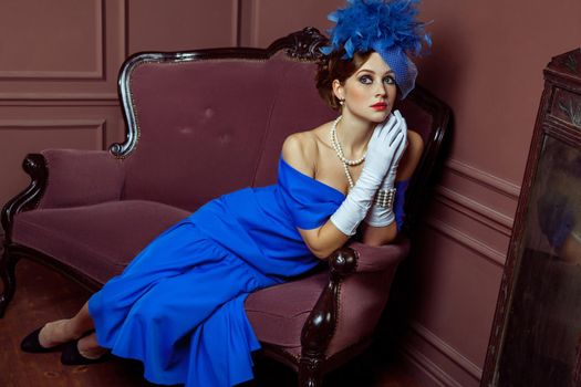 Old Fashioned retro great britain style photography. Beautiful young caucasian model in blue dress and fashion makeup and hat posing sitting on the artificial sofa and looking into the distance.