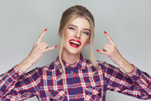 funny kissing beautiful blonde girl with pink checkered shirt, collected updo hairstyle and makeup standing and showing Ok sign and looking at camera with wink. indoor , isolated on gray background.