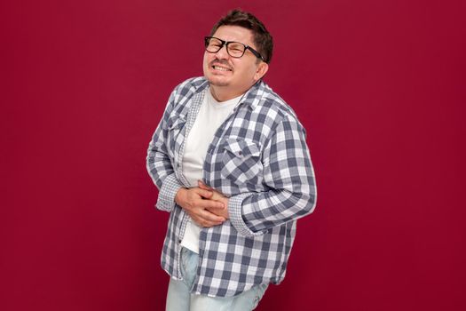 Stomach pain. Portrait of middle aged man in casual checkered shirt and eyeglasses standing, feeling bad and holding his painful sick belly. indoor studio shot, isolated on dark red background.
