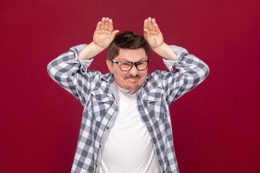 Portrait of funny handsome middle aged business man in casual checkered shirt and eyeglasses standing with bunny ears gesture on head, looking at camera. studio shot, isolated on dark red background.