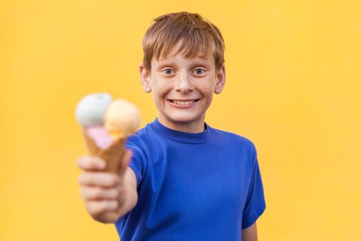 Blonde beautiful boy with freckles and blue T-shirt showing ice cream and looking at camera with big eyes and toothy smile. Studio shot, isolated on a yellow background.