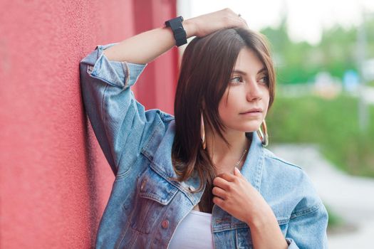 Portrait of beautiful brunette young woman with makeup in denim casual style standing, posing with hand on head and looking away. outdoor shot near red wall in summertime.