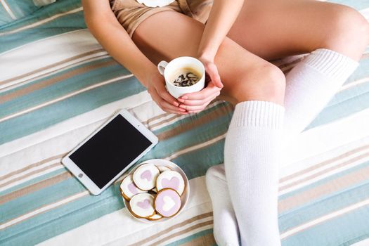 Woman using tablet at cozy home atmosphere on the bed. Young woman with cup of cocoa or coffee in hands enjoying free time with comfort. Soft light and comfy lifestyle concept. top view point..