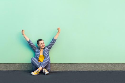 Portrait of happy rejoicing handsome beautiful short hair young woman in casual striped suit sitting and celebrating her victory or success. indoor studio shot isolated on light green background.