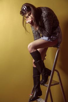 Beautiful young asian model in blue denim skirt and black blouse and fashion makeup posing sitting on stepladder and looking at camera. Studio shot. High-fashion photo.