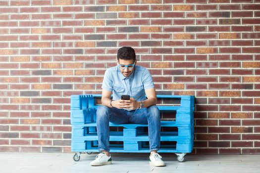 Full length portrait of successful handsome young bearded man in casual style, eyeglasses sitting on blue wooden pallet, holding, looking at mobile smartphone. indoor studio shot on brown brick wall.