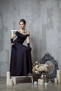 Beautiful young fashion caucasian model in black long dress and fashion makeup and beautiful hairdo posing sitting on the chair and looking at camera. Studio shot.