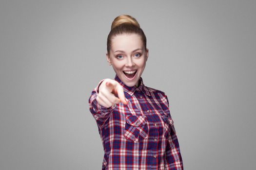 Amazed beautiful blonde girl with pink checkered shirt, collected hairstyle and makeup standing, looking at camera and with finger pointing and shocked. indoor studio shot, isolated on gray background