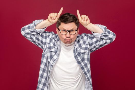 Portrait of angry middle aged business man in casual checkered shirt and eyeglasses standing with cow gesture horns on head and looking at camera. indoor studio shot, isolated on dark red background.
