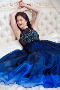 Attractive brunette woman with beautiful makeup in blue evening dress posing while sitting on bed and looking at camera with toothy smile. indoor studio shot.