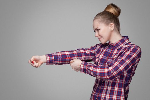 profile side view of blonde girl in red, pink checkered shirt, collected bun hairstyle standing and fists with srious face or try to pulling. indoor studio shot. isolated on gray background